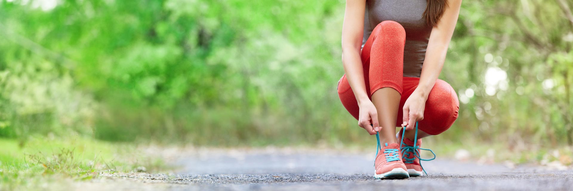 This is an image of a woman on an outdoor trail who has bent down to tie her bright red running shoe.