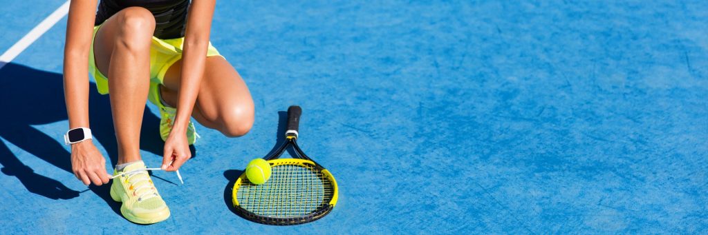 This is an image of a woman on a bright blue tennis court who has bent down to tie her bright yellow tennis shoe. There is a yellow racket and tennis ball beside her.