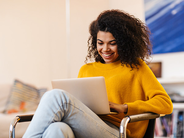 Happy young woman in a yellow sweater seated in a chair using a laptop computer.