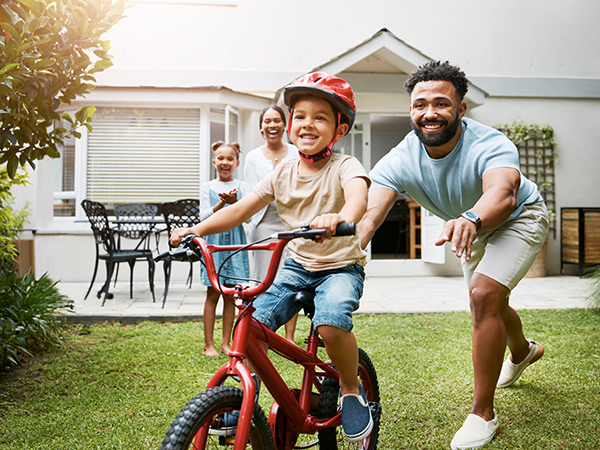 Happy smiling family of four in a front yard teaching a child in a helmet to ride a bicycle.