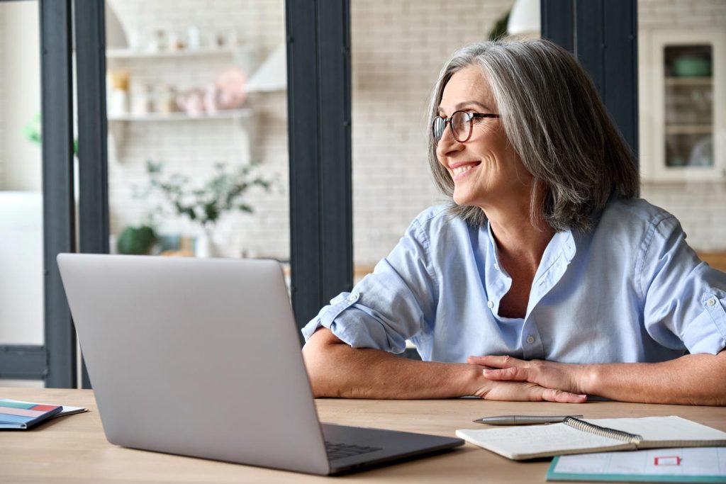 This is an image of a smiling middle-aged woman with glasses at a desk using a laptop computer.